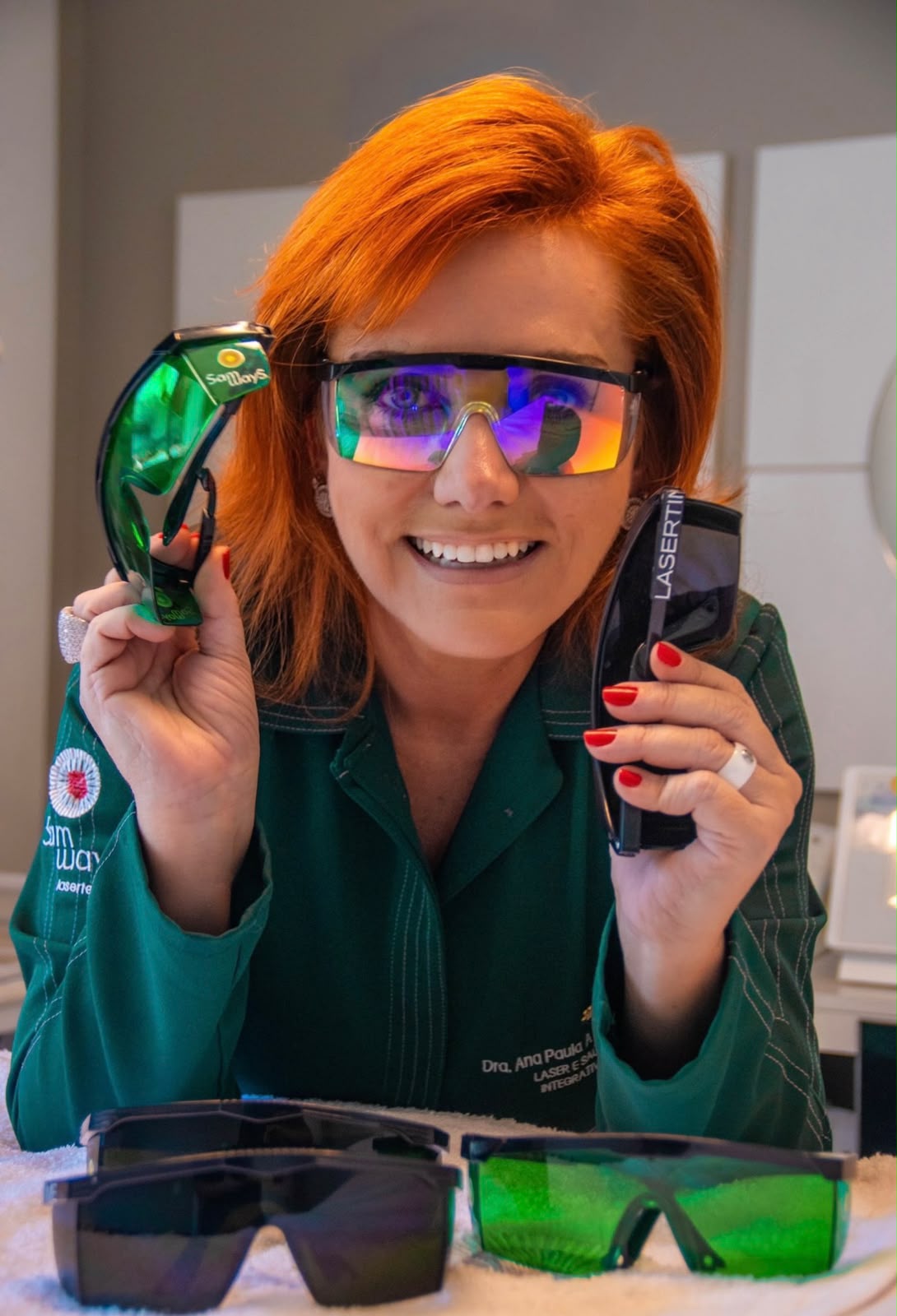 Dra. Ana Paula Samways, de jaleco verde, segurando dois óculos de proteção para laser em sua clínica.